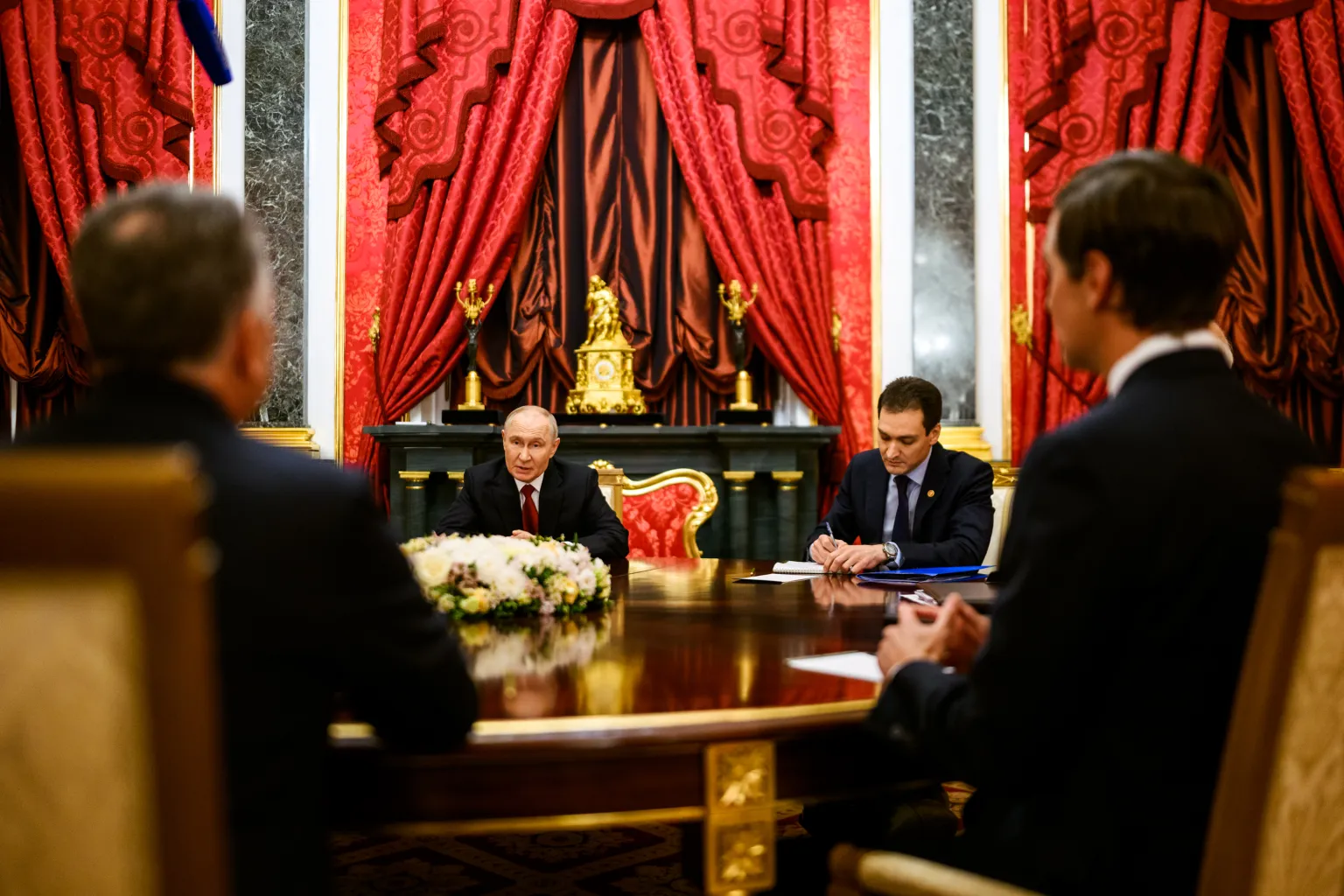 U.S. envoys, seen from behind, at a table facing the Russian side in an ornate hall with red drapes and a gilded clock