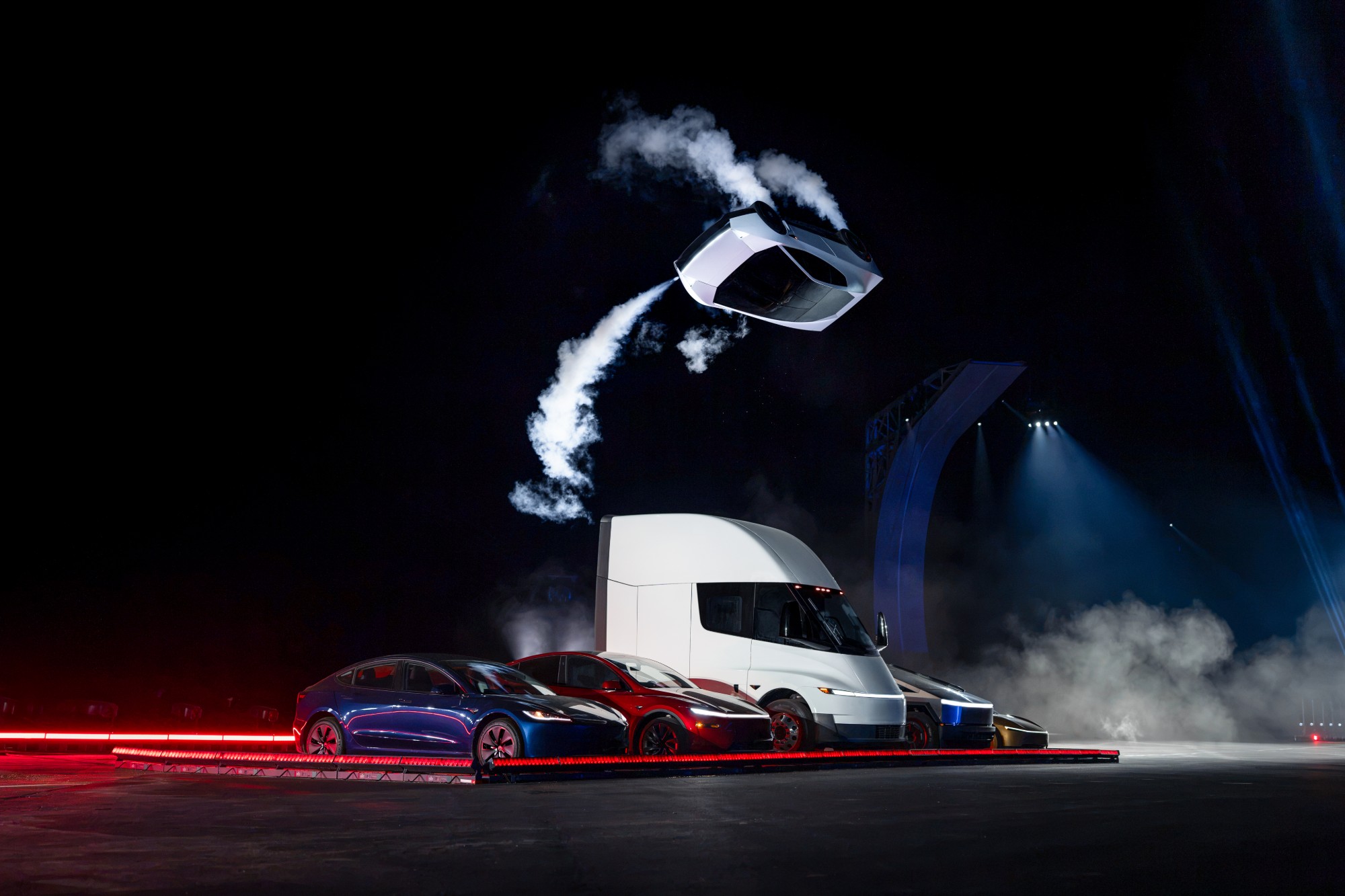 A white Tesla Roadster flips through the air trailing white gas plumes above a line-up of Tesla cars, a Semi truck and a Cybertruck on a dark stage