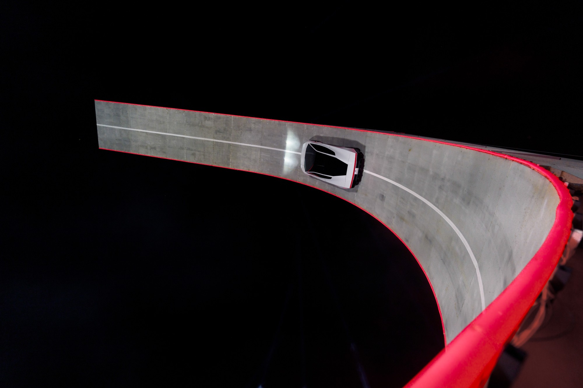 Overhead view of the white Roadster driving up a steep, curved ramp lined in red, the track turning towards vertical