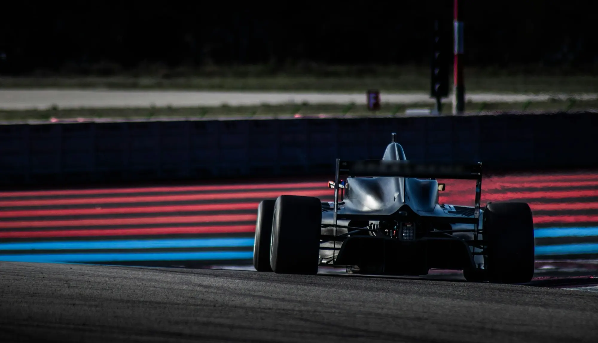 A single-seater race car seen from behind at dusk, passing red and blue kerbs